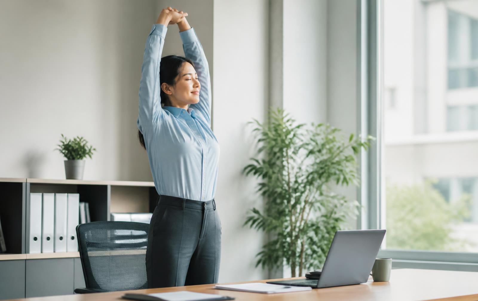 An office worker standing beside a desk and stretching their arms overhead during a break