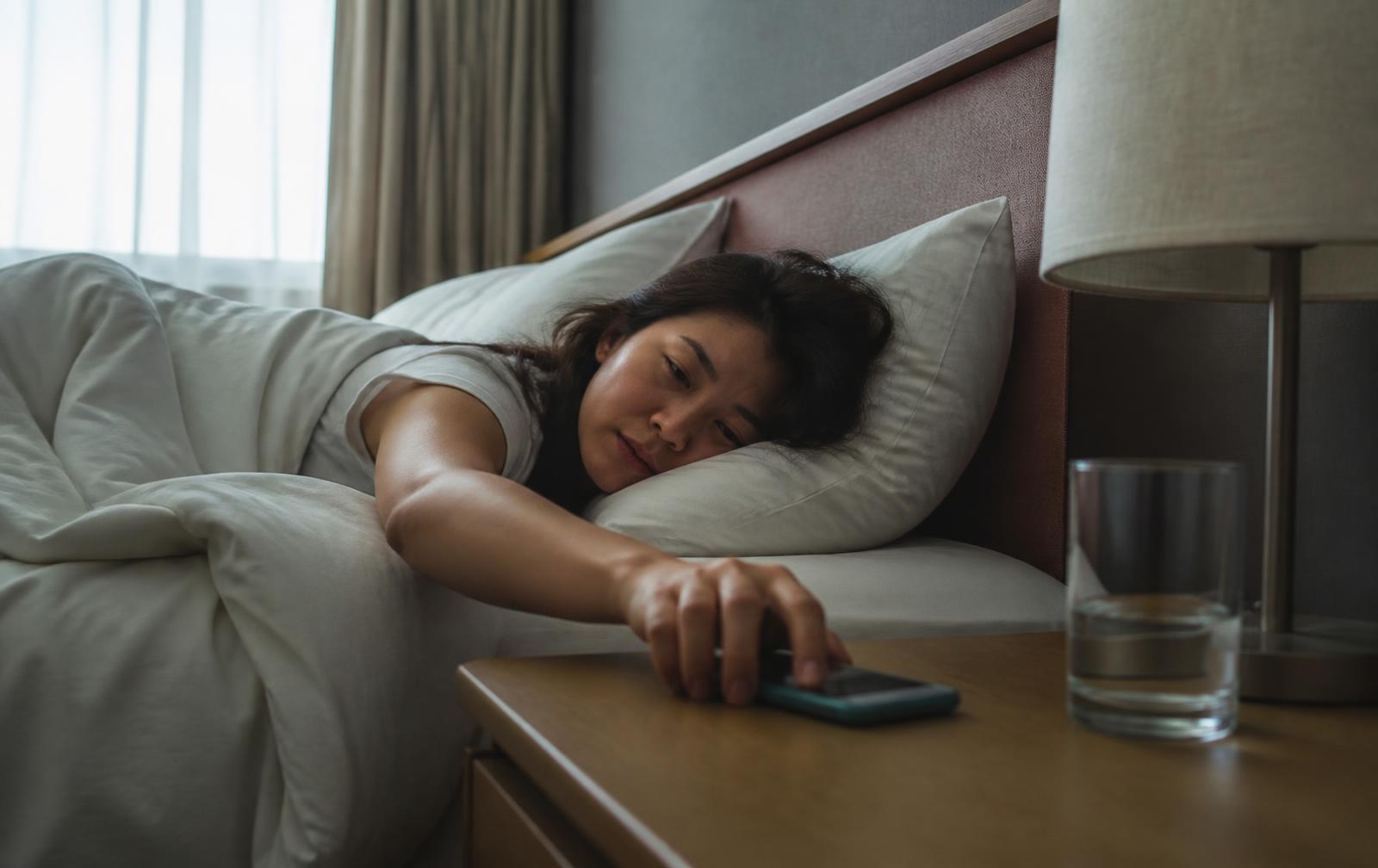 A person in bed reaching for a smartphone on the nightstand in the early morning