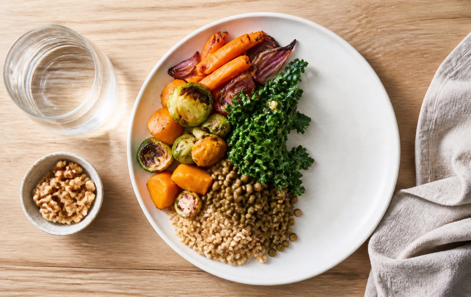 Overhead view of a plate half filled with roasted vegetables, kale, lentils and whole grains beside a glass of water and a bowl of walnuts