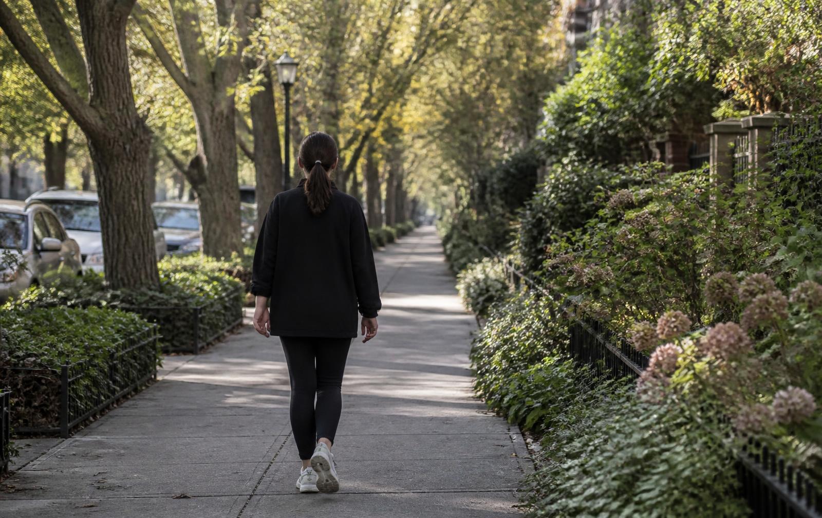 A person walking alone along a tree-lined neighborhood sidewalk in afternoon sunlight